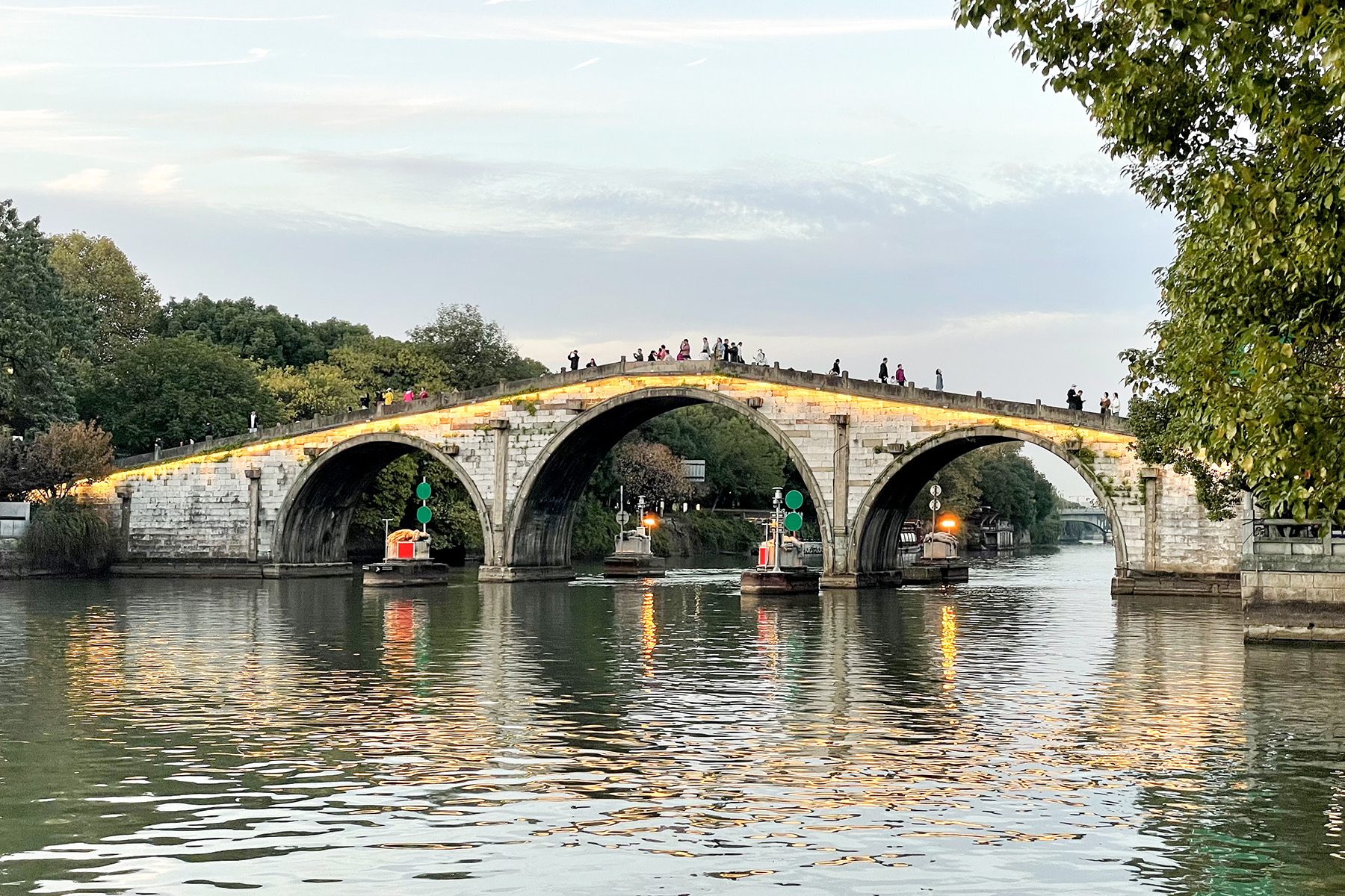 Hangzhou's Gongchen Bridge lit at dusk, its three stone arches reflected in the Grand Canal with navigation lights on the water