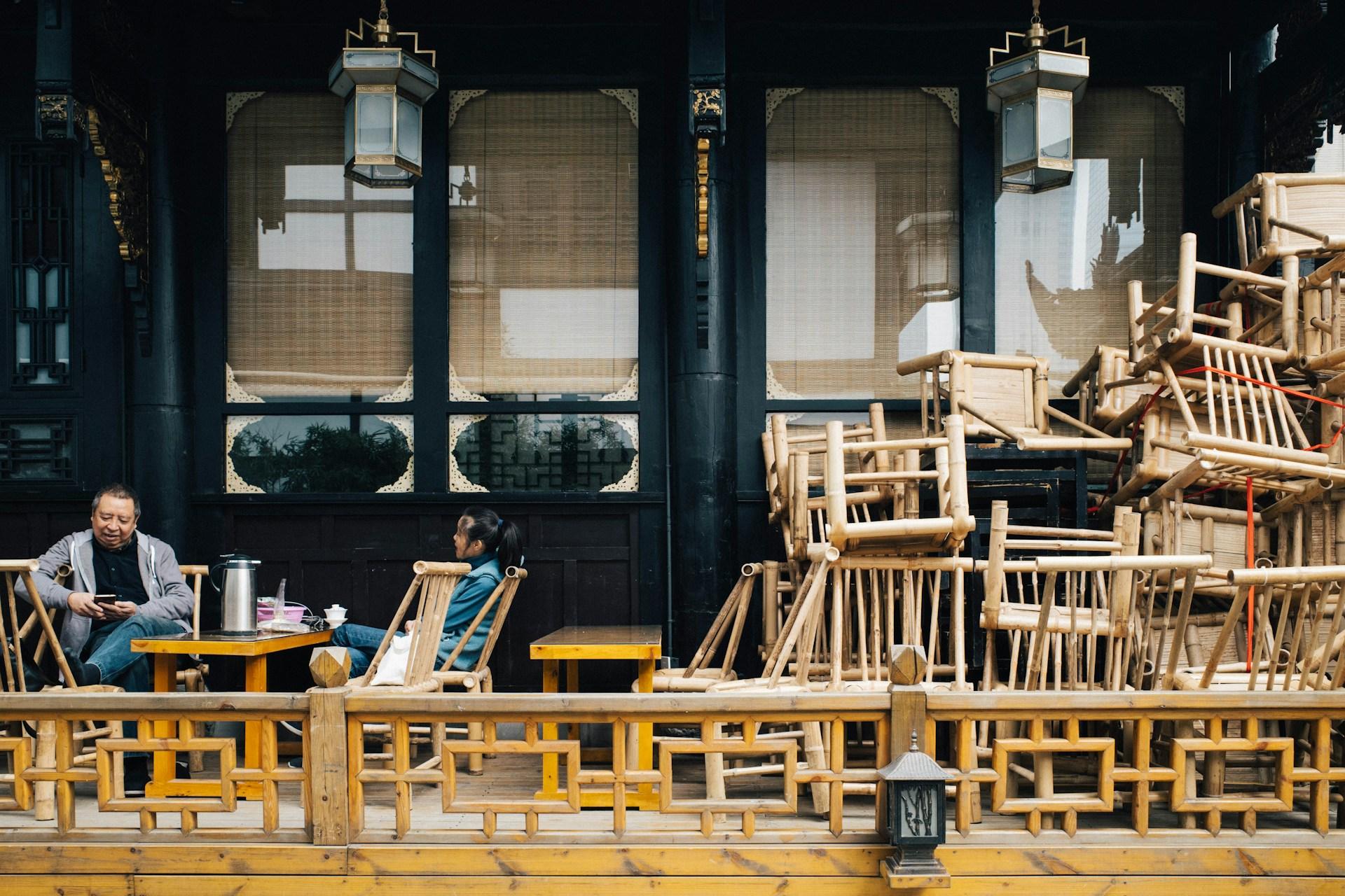 Two regulars drinking tea among stacked bamboo chairs at an old Chengdu tea house