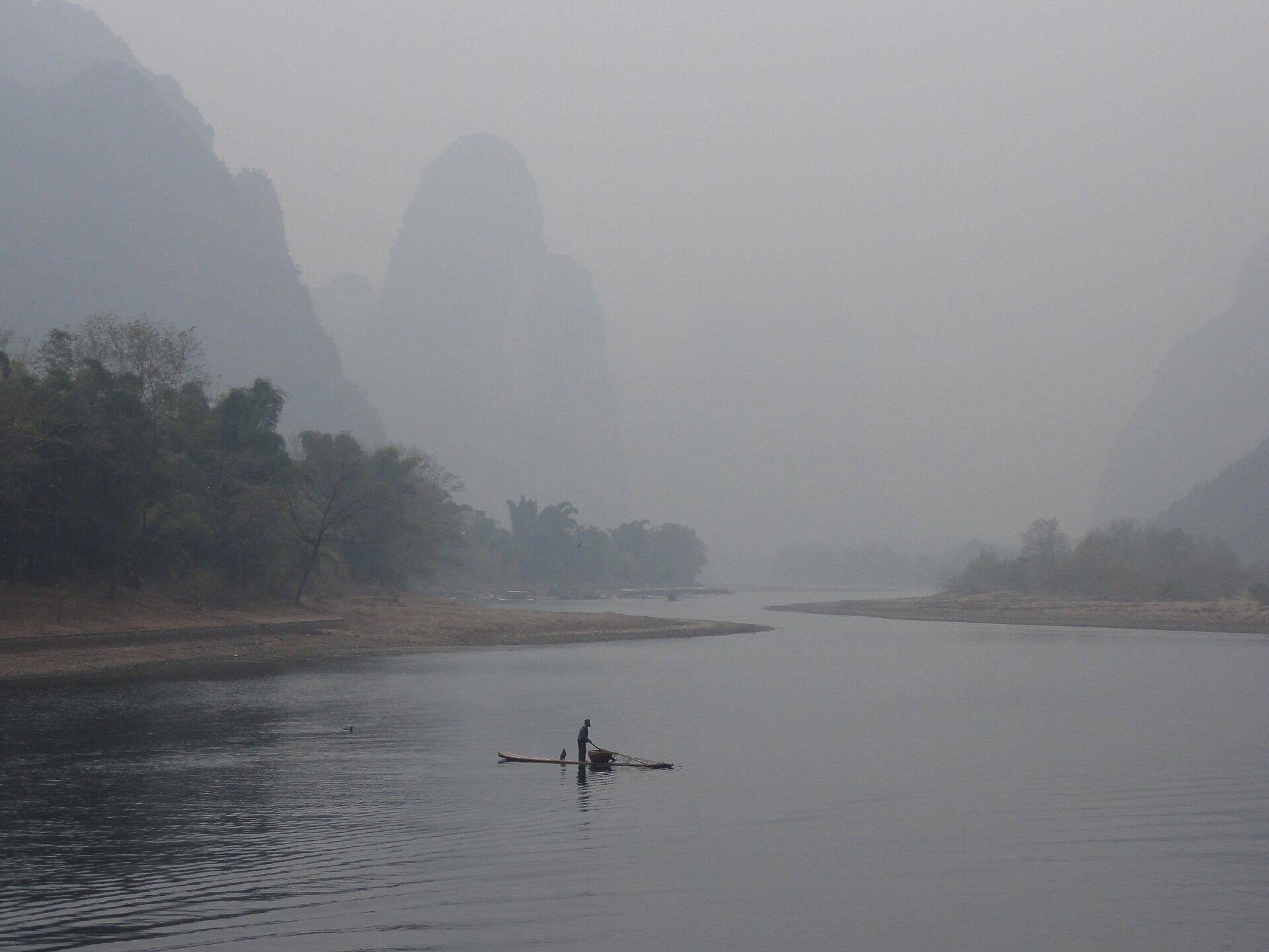 A lone cormorant fisherman poling a bamboo raft through dense morning mist on the Li River, karst peaks dissolving behind
