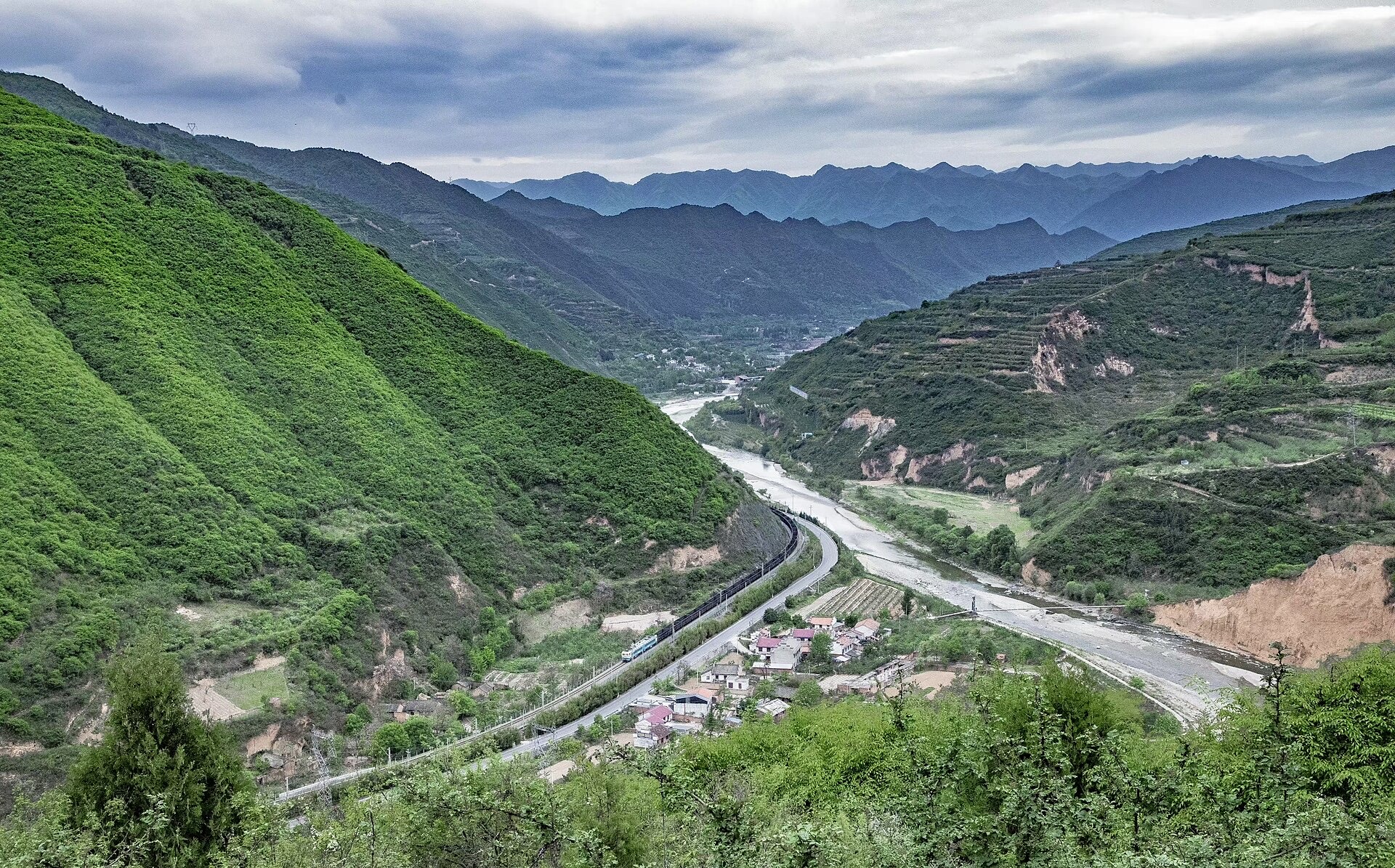 A long freight train winding through a deep green valley in the Qinling mountains
